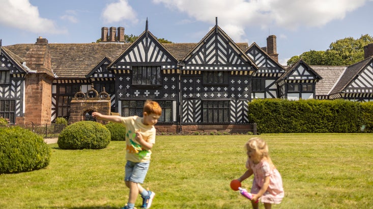 A girl and a boy play on the lawn in front of the black and white Tudor house at Speke Hall.
